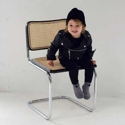 Criança sentada em cadeira moderna, vestindo casaco de couro e meias Attipas Koala Grey, com um estilo urbano.