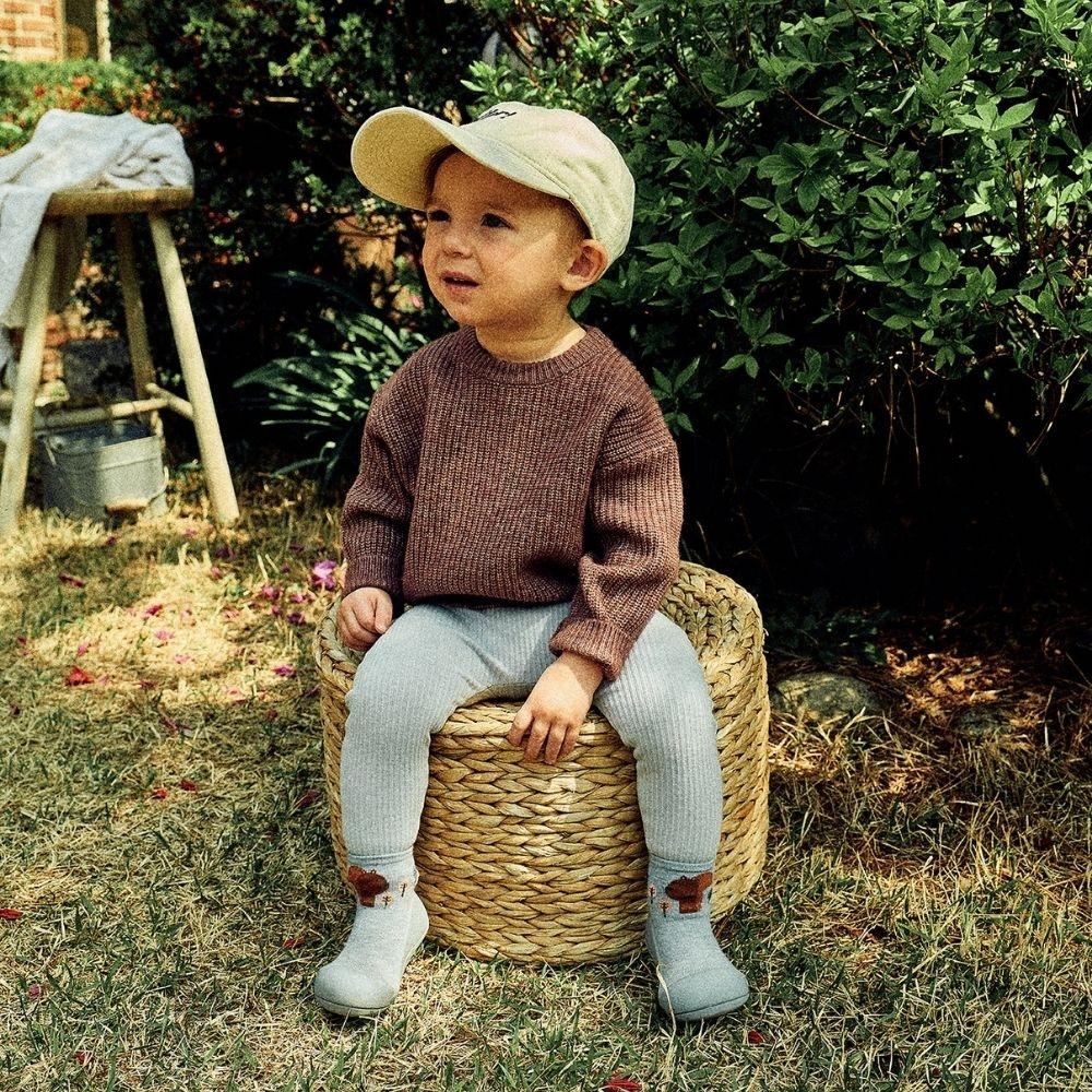 Criança sentada em um cesto, vestindo suéter castanho e calças cinzentas, com boné claro, em ambiente natural.