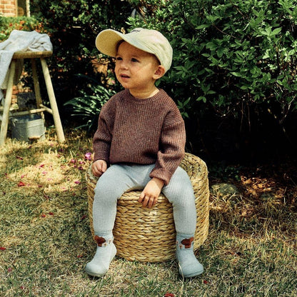 Criança sentada em um cesto, vestindo suéter castanho e calças cinzentas, com boné claro, em ambiente natural.