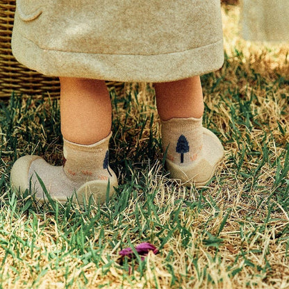 Pequenos pés com meias de árvore, calçando Attipas Woodland Beige Fox, sobre a relva numa tarde ensolarada.