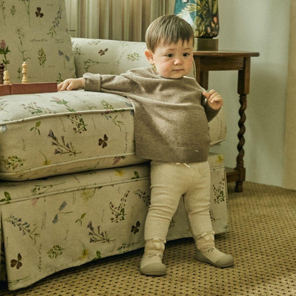 Bebé em pé ao lado de um sofá floral, vestindo suéter e calças, com meias Attipas Rabbit Beige.